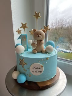 a blue cake with gold stars and a teddy bear on top is sitting in front of a window