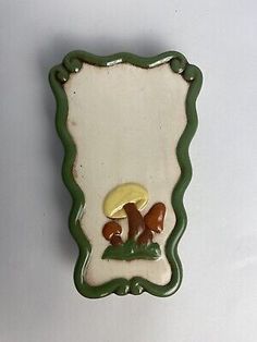 a green and white ceramic wall hanging with mushrooms on the bottom, in an ornate frame