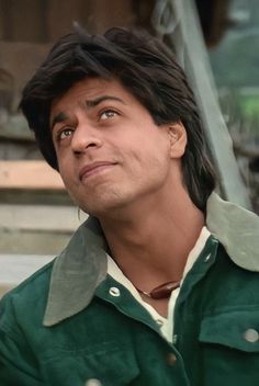 a man with black hair and green shirt looking off to the side in front of a building