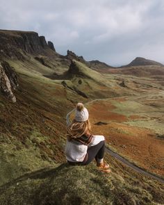 a woman sitting on top of a grass covered hill next to a lush green hillside