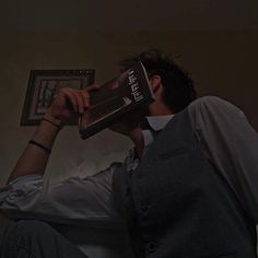 a man sitting on a couch while holding a book up to his face with both hands