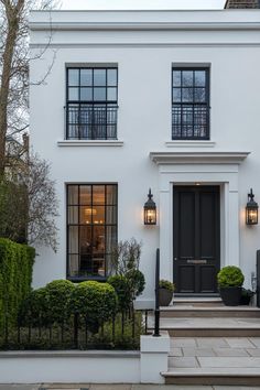 a white house with black doors and windows on the front steps, surrounded by hedges