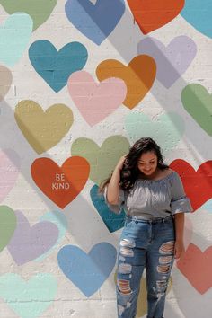 a woman standing in front of a wall with hearts painted on it and the words be kind