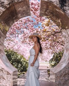 a woman wearing a white dress and hat standing in front of a tree with pink flowers