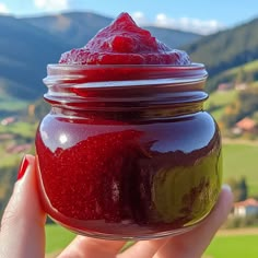 a hand holding a jar filled with red liquid on top of a lush green hillside