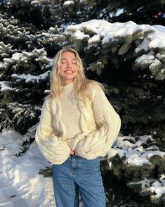 a woman standing in front of snow covered evergreen trees wearing a white sweater and jeans