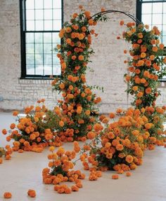 an arrangement of orange flowers and greenery in front of a brick wall with large windows