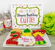 decorated cookies and watermelon are sitting on a plate next to a sign that says two - it - fruit happy birthday cutie