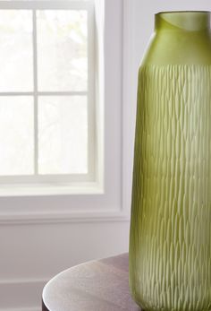 a green vase sitting on top of a wooden table next to a window with white curtains