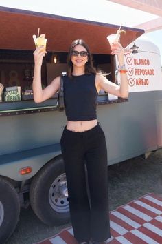 a woman standing in front of a food truck holding up two drinks and posing for the camera