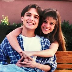 a young man and woman sitting on a wooden bench smiling at the camera with their arms around each other