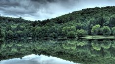 a large body of water surrounded by trees and mountains in the background with dark clouds