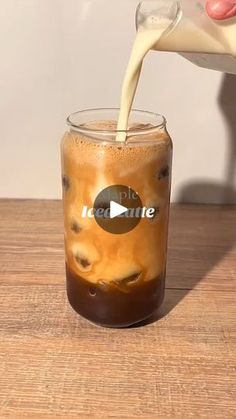 a person pouring milk into a glass filled with iced coffee on top of a wooden table