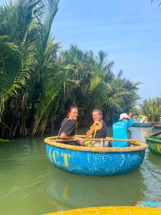 two people in a blue and yellow boat on the water with palm trees behind them