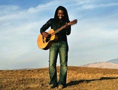 a man standing on top of a dry grass covered field holding a guitar in his hands