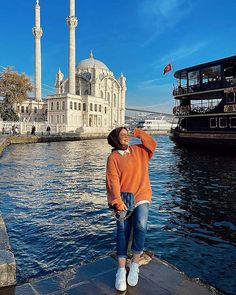 a woman in an orange sweater is standing on the edge of a dock with her arms outstretched