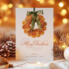 a christmas card with a wreath on it and pine cones in the foreground, surrounded by lights