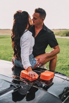 a man and woman kissing on the hood of a car in front of a grassy field