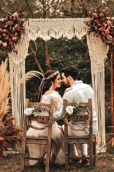 a bride and groom sitting on wooden chairs in front of an outdoor wedding ceremony arch