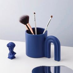 a blue cup with brushes in it sitting next to a mirror and a small figurine