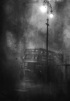 a double decker bus parked next to a street light on a foggy night in black and white