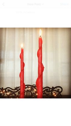 two red candles sitting on top of a table next to a window with white curtains