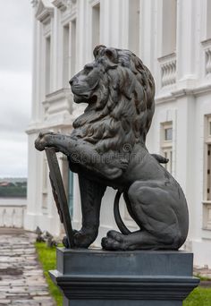 a statue of a lion holding a shield in front of a white building with green grass and cobblestones