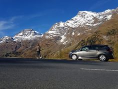 a person standing next to a car on the side of a road with mountains in the background