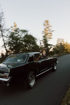 an old black car driving down the road with flowers in the back seat and on the passenger seat