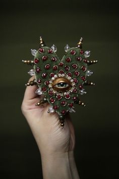 a hand holding a green brooch with red and white beads on it's face