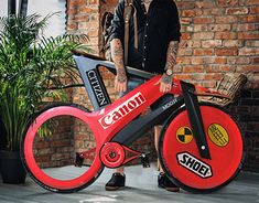 a man standing next to a red bike with a giant wheel on it's side