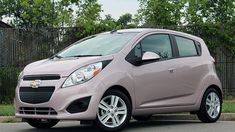 a pink chevrolet spark is parked on the side of the road in front of a fence