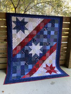 an american flag quilt hanging on a wooden bench in front of a tree and fence