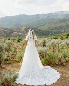 a woman in a wedding dress looking out at the mountains with flowers on her back