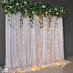 white flowers and greenery are hanging on the sheer curtain with lights in front of them