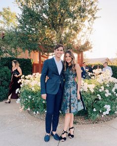 a man and woman standing next to each other in front of some bushes with white flowers