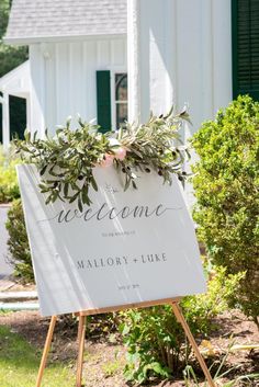 a welcome sign in front of a white house with greenery on the top and bottom