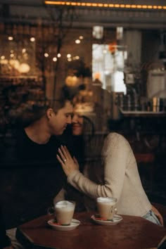 two people sitting at a table with cups in front of them and one person touching the other's face