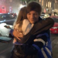 a man and woman hugging on the street at night time with cars in the background