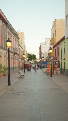 people are walking down the street in front of buildings and benches with lights on them