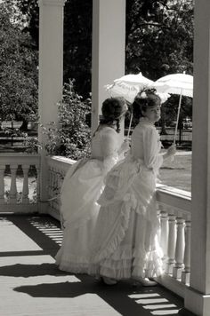 two women in dresses are standing under an umbrella on the porch with their backs to each other