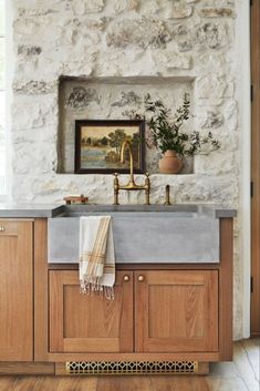 a kitchen sink sitting under a window next to a wooden cabinet and counter top in front of a stone wall