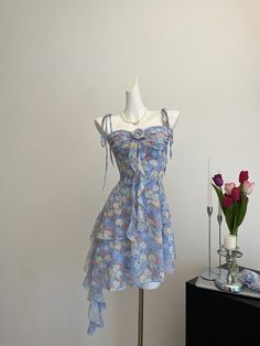 Circuitous and crafted with care, this blue floral dress weaves its way through timeless fashion with a modern twist. The layers of lightweight fabric create a gentle cascade, giving this dress a buoyant, ethereal quality that’s perfect for spring and summer events. Decorated with a vibrant array of floral patterns in soft hues, the dress features dainty shoulder straps and a rosette detail for a touch of romance. Its asymmetrical hemline and flowing silhouette ensure it moves gracefully with th Light Blue Dress-down Dresses For Summer, Chiffon Ruffled Dress For Spring Garden Party, Spring Chiffon Dress With Ruffles For Garden Party, Spring Chiffon Dress With Ruffles, Spring Chiffon Dress With Ruffles For Casual Wear, Spring Casual Chiffon Dress With Ruffles, Ruffled Chiffon Dress For Spring Garden Party, Light Blue Dresses For Summer Dress Down, Spring Chiffon Tiered Dress