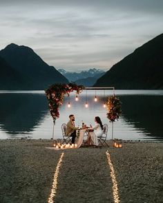 two people sitting at a table with candles in front of them on the shore of a lake