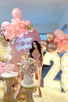 a woman standing in front of a birthday cake with balloons and flowers on the table