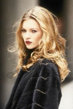 a woman with long blonde hair is walking down the street wearing a black coat and red lipstick