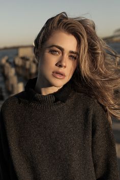 a woman with long hair standing by the water wearing a sweater and looking at the camera