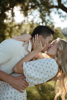 a man and woman kissing each other while standing in front of some trees on a sunny day