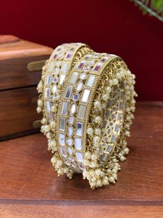 This authentic Kalinda bangle is a classic blend of traditional aesthetics and modern craftsmanship. Intricately crafted of brass with real mirror and pearl work, its one-inch width and champagne stone sparkle make it a chic, timeless addition to any ensemble. Comes in a pair. Elegant Festive Cutdana Bangle, Elegant Kundan Bangle With Stone Work, Elegant Gota Work Bangle As Gift, Elegant Gota Work Bangle For Gifts, Celebration Bangle With Gota Work, Elegant Wedding Bangle With Gota Work, Elegant Bangle With Gota Work For Festivals, Festive Silver Bracelets With Gota Work, Elegant Festival Bangle With Gota Work