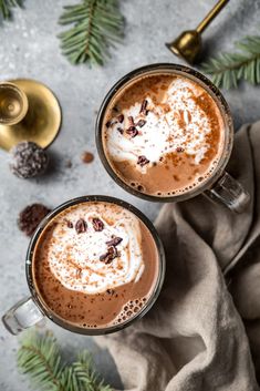 two cups of hot chocolate with whipped cream on top and pine cones in the background